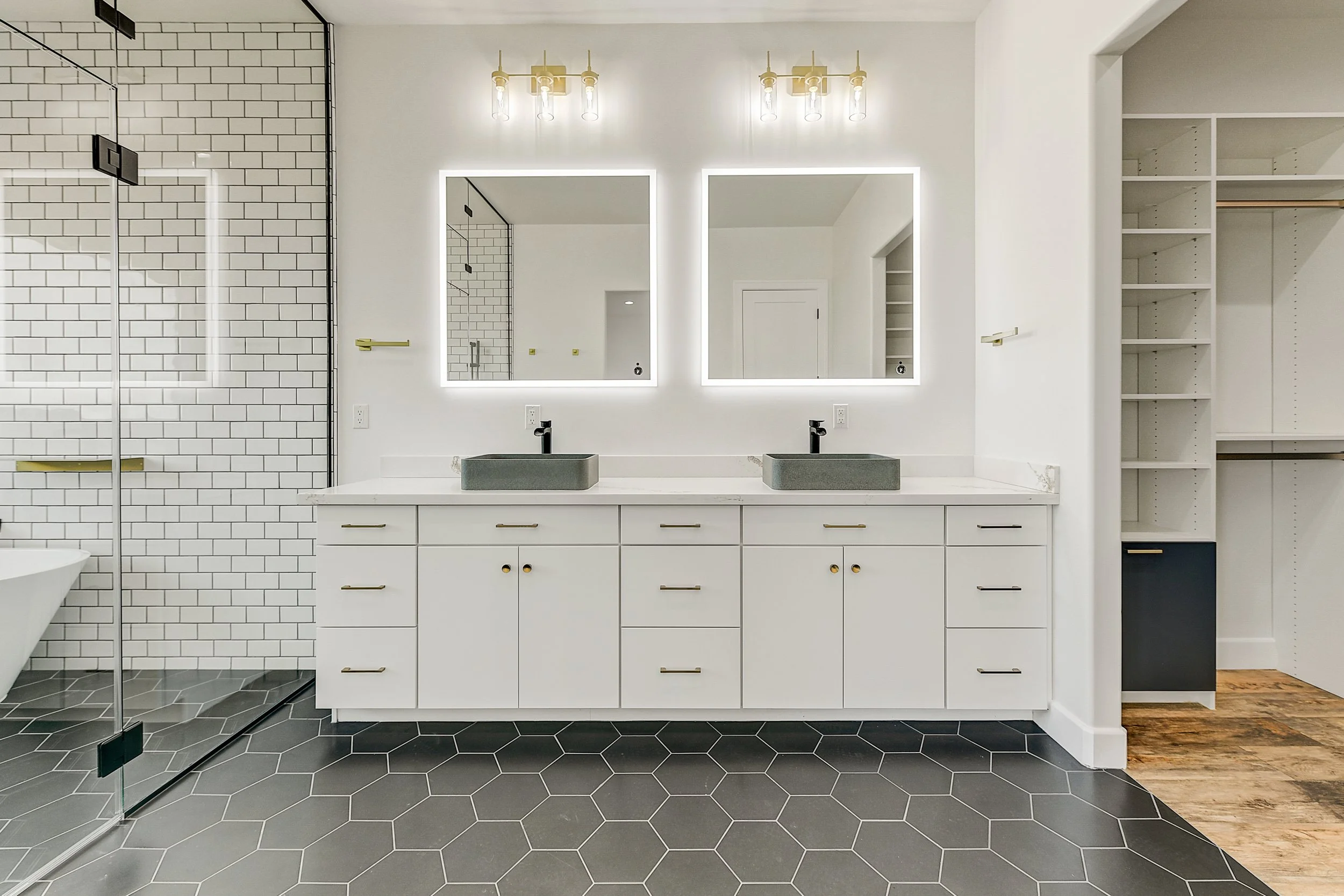 Modern bathroom with dual sinks, illuminated mirrors, white cabinetry, black and gold fixtures, hexagonal black floor tiles, and a walk-in shower with white subway tiles and black framing.
